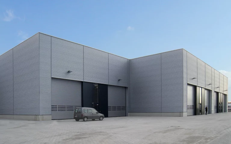 Neubau Siemens Multifunktionshalle Wegberg Außenaufnahme mit hinterlüfteter Trapezblechfassade, Betonsockel, Portalen, Sektionaltoren und Umfahrt.