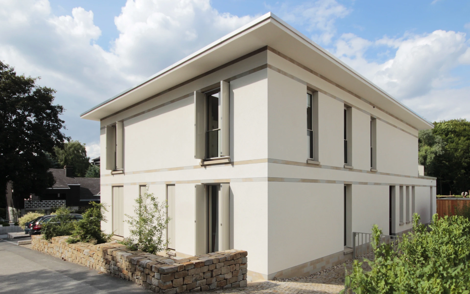 Neubau Haus D in Bochum Ansicht von der Straße mit Putzfassade mit Faltschiebeläden, eingelegten Bändern und Sockel sowie bepflanzter Bruchsteinmauer aus Ruhrsandstein.