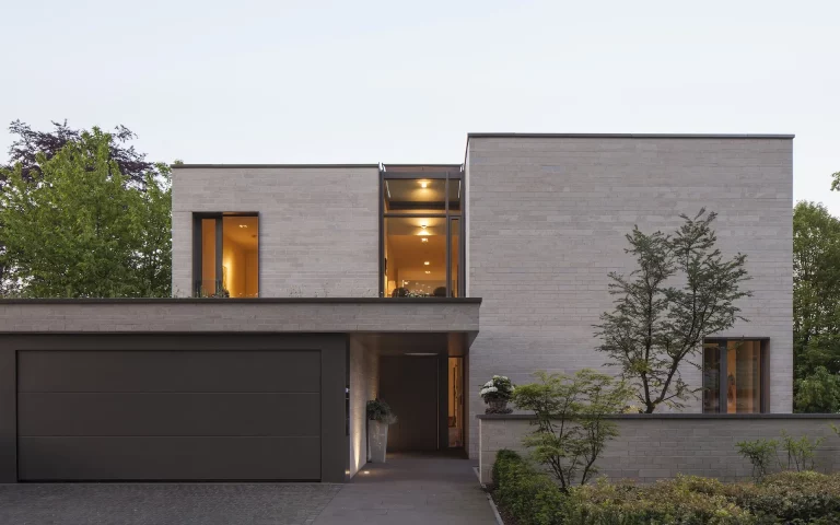 Neubau Einfamilienhaus S in Essen mit Eingangssituation, Vorfahrt, Natursteinfassade, raumhohen Verglasungen, Gartenmauer und Bäumen.