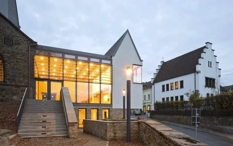 Neubau Gemeindezentrum St. Viktor Ansicht vom Kirchgarten mit Blick auf raumhoch verglasten Gemeindesaal, Giebel und Satteldach, Natursteinsockel und Anschluss Kirche.