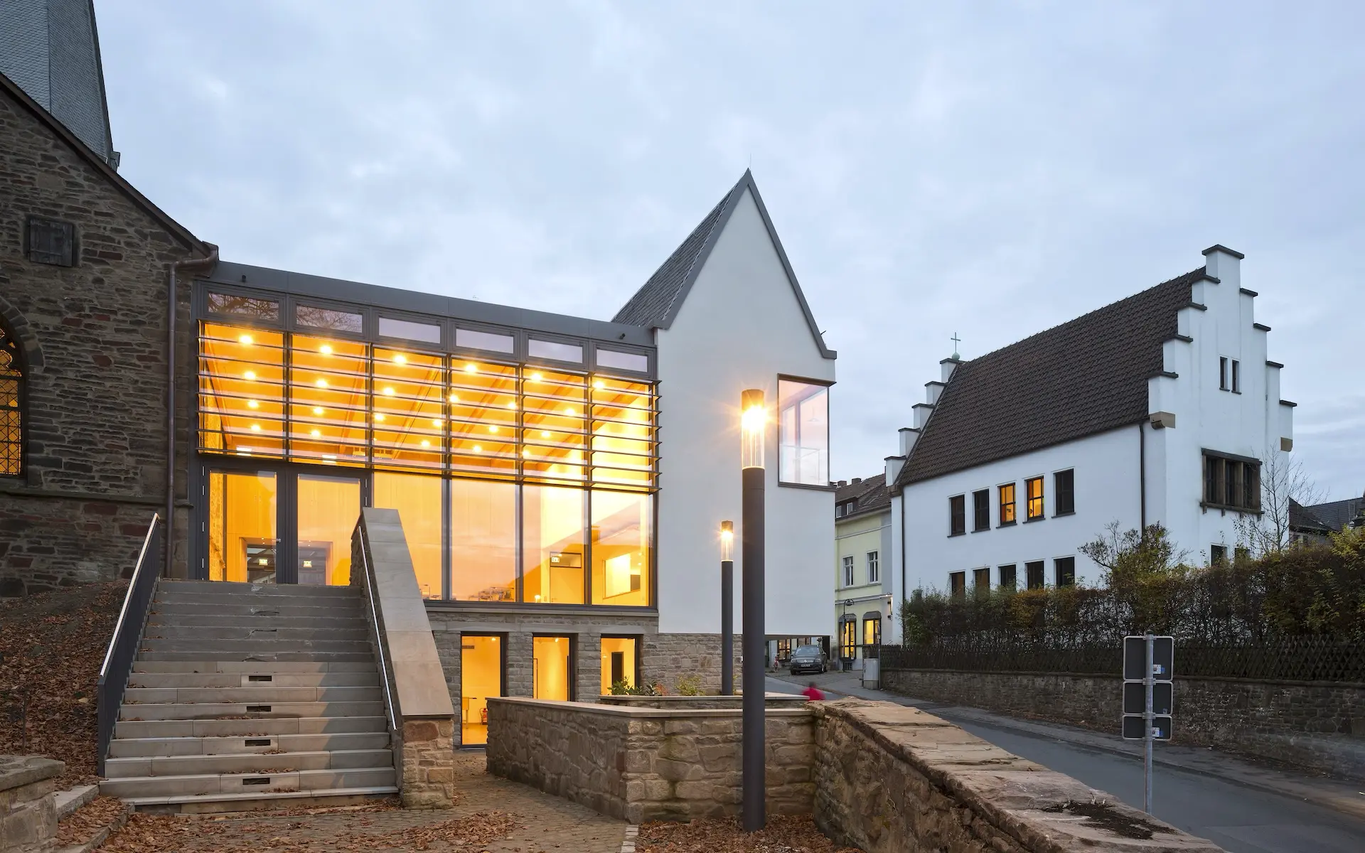 Neubau Gemeindezentrum St. Viktor Ansicht vom Kirchgarten mit Blick auf raumhoch verglasten Gemeindesaal, Giebel und Satteldach, Natursteinsockel und Anschluss Kirche.