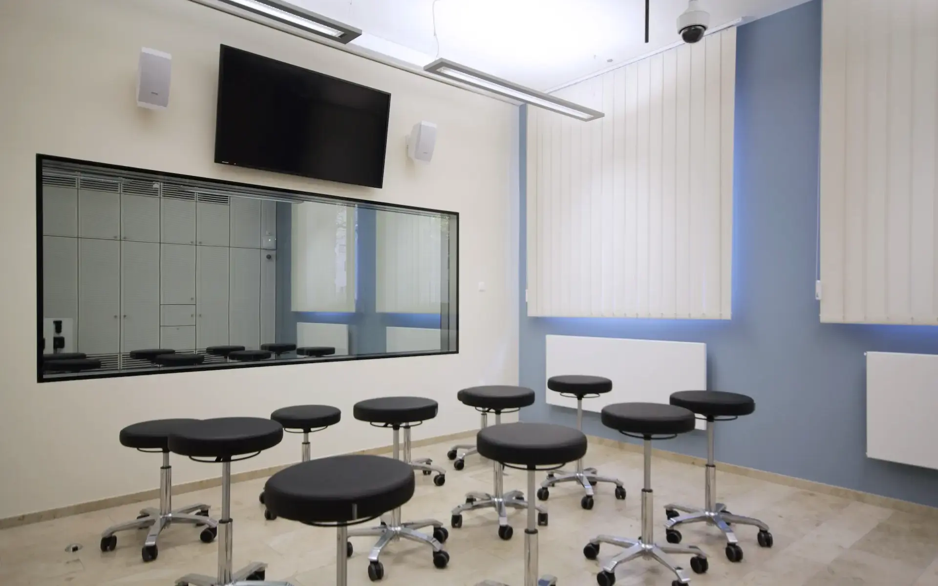 Das Bild zeigt den Neubau des Lehr- und Lernzentrum in Essen. Abgelichtet ist ein Seminarraum, welcher durch eine einseitige Glasscheibe den Blick in einem Simulationsraum erlaubt. Hier werden praxisnahe Situation im Klinikalltag nachgestellt und können durch diesen Seminarraum zeitgleich besprochen und evaluiert werden.