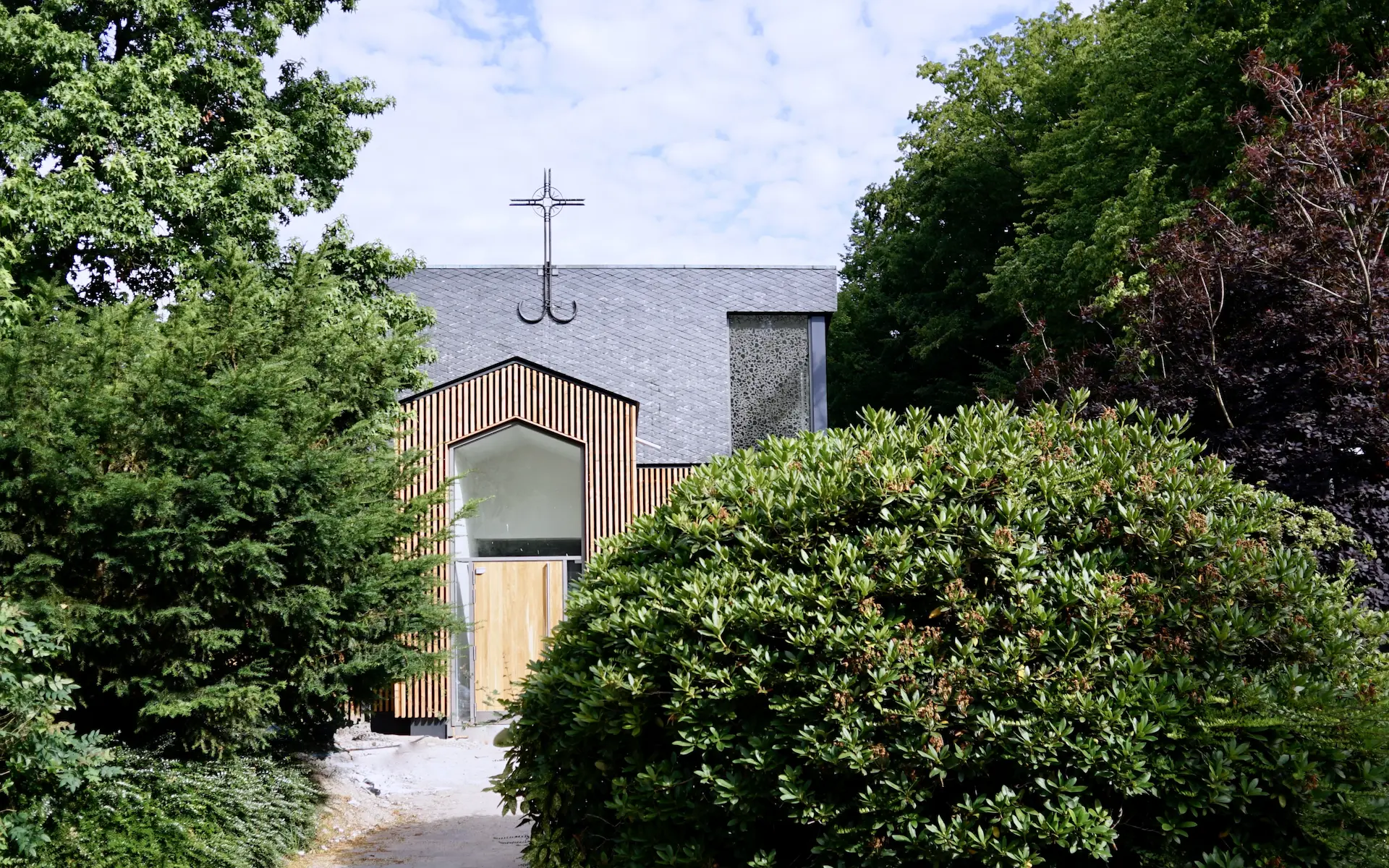 Umbau St. Altfrid in Essen Aussenansicht vom Vorplatz mit Buschwerk im Vordergrund, Kircheneingang, Kirchenkreuz, Kirchenfenster und Eichentür.