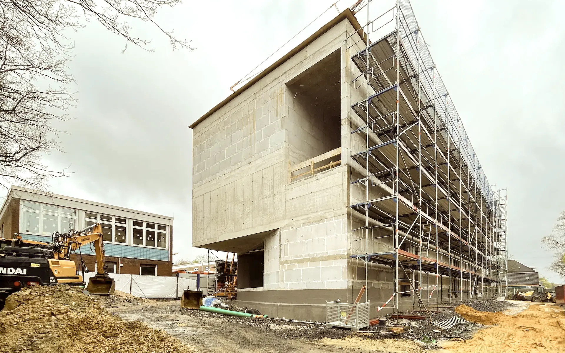 Erweiterung und Sanierung Hövelschule Essen Baustellenansicht im Rohbau mit Betonwänden und Unterzügen, zweigeschossigen Fensteröffnungen, Attika und Bagger.