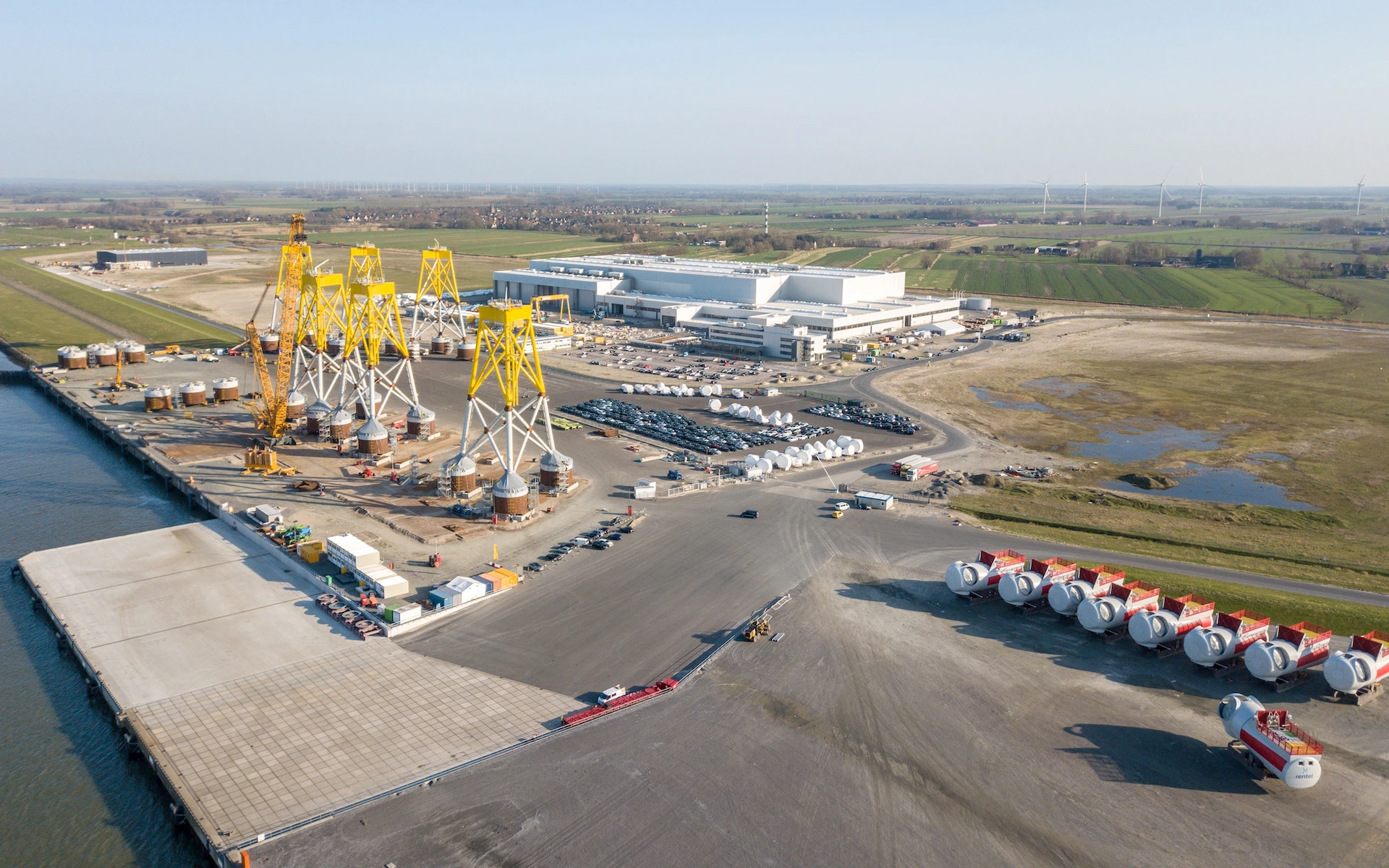 Neubau Siemens Windturbinen Fabrik in Cuxhaven als Vogelperspektive mit RoRo-Rampe, Windturbinen, Lagerflächen, Büros und abgestaffelten Industriebau mit Produktionslinien.