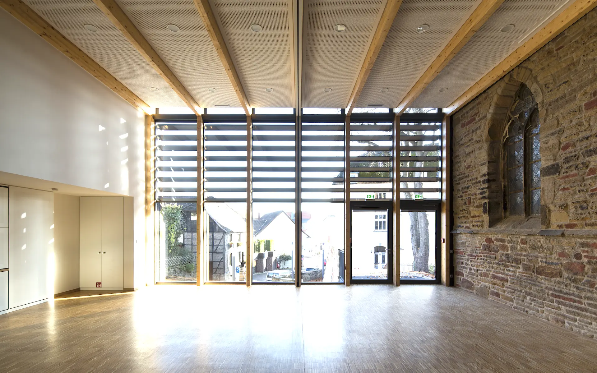 Neubau Gemeindezentrum St. Viktor Innenraum des Gemeindesaals mit raumhoher Verglasung, Holztragwerk, Anschluss an Kirchenmauerwerk, Parkettboden und Küchenausgabe.