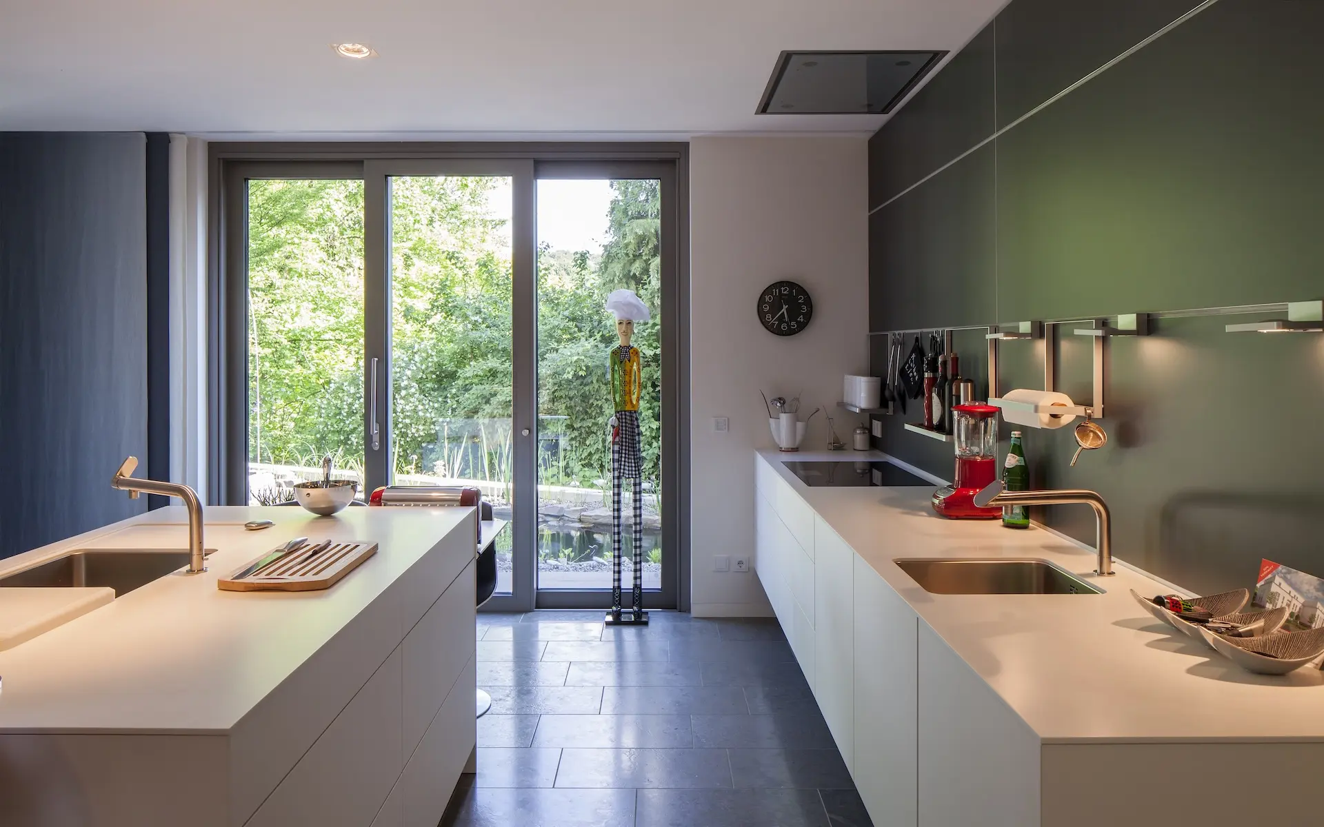 Neubau Einfamilienhaus S in Essen Küche mit raumhohen Fenstern, freistehendem Küchenblock, grüner Wandtäfelung und Natursteinboden.