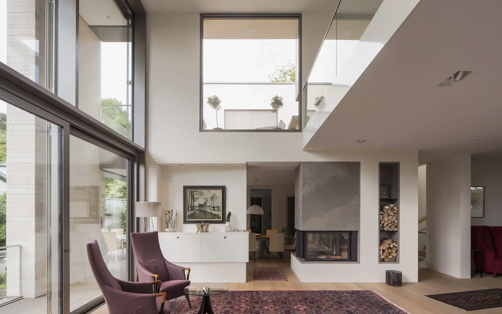 Neubau Einfamilienhaus S in Essen mit einer Innenraumaufnahme des Wohnzimmers mit gläserner Galeriebrüstung, Kamin, zweigeschossigen Luftraum und Glasfassade.