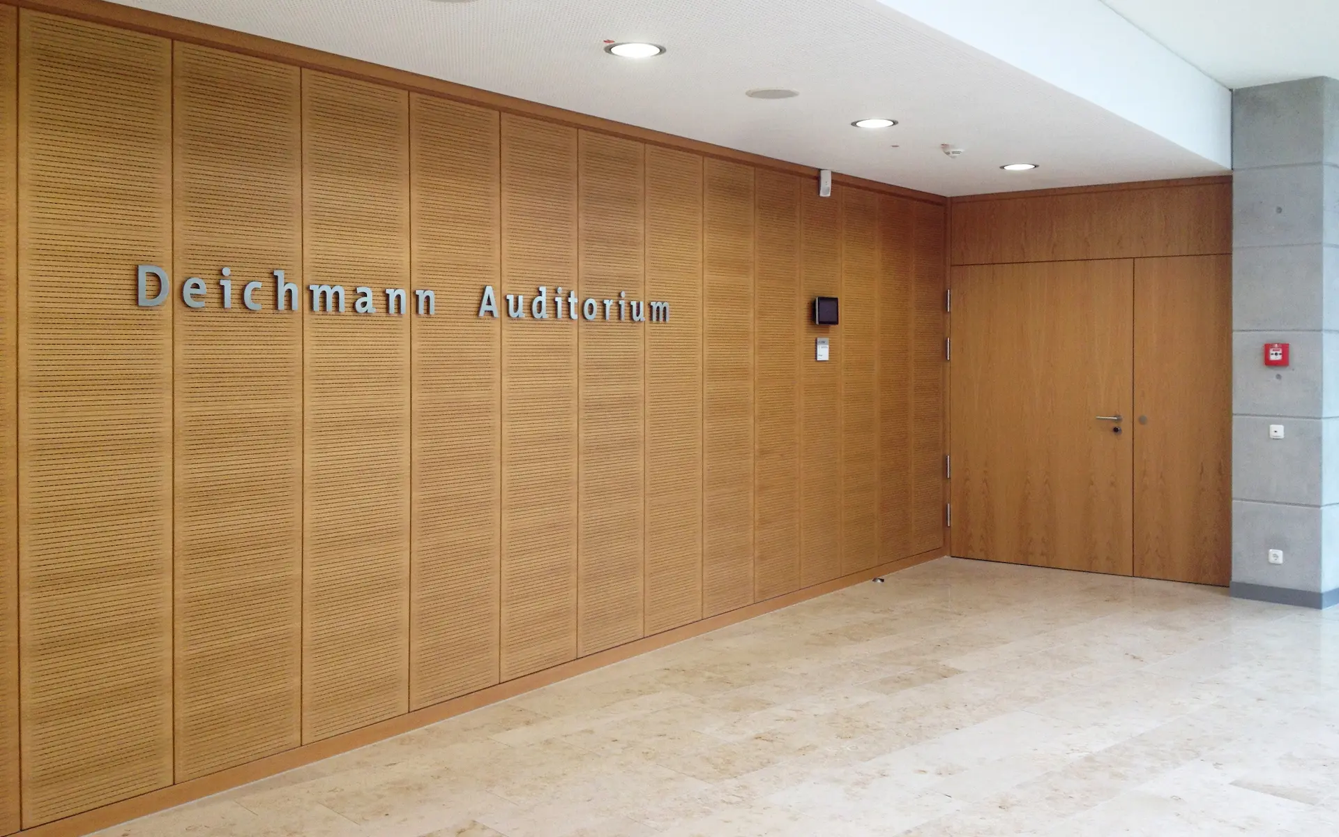 Neubau Lehr- und Lernzentrum des Universitätsklinikums Essen Hörsaalfoyer mit Holztüren und Holzverkleidungen, Downlights, Sichtbetonwänden und Natursteinboden.