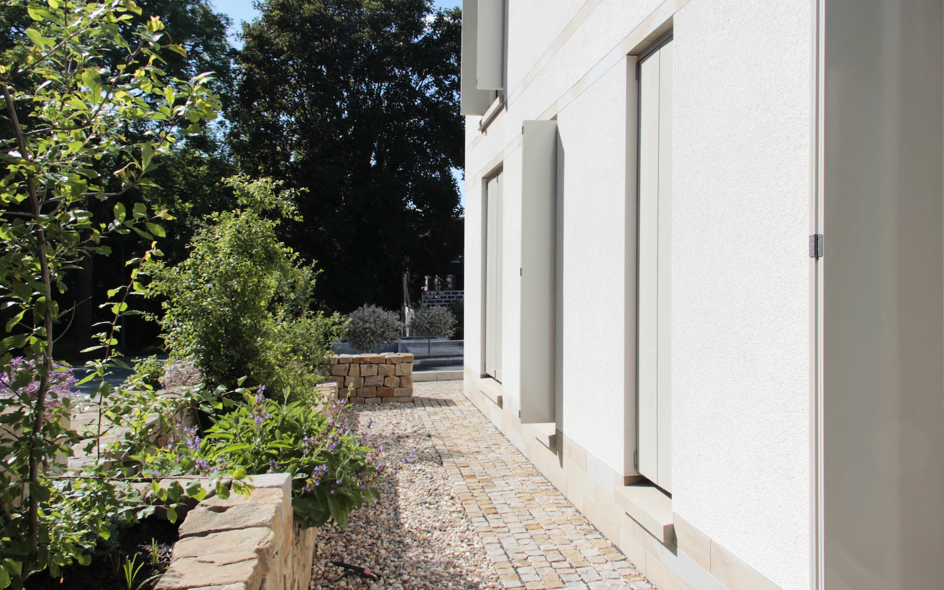 Neubau Haus D in Bochum Detail der Putzfassade mit Faltschiebeläden, der Sohlbänke, des Sockels, der bepflanzten Bruchsteinmauer sowie des Pflasters aus Ruhrsandstein mit unterschiedlichen Oberflächenbearbeitungen.