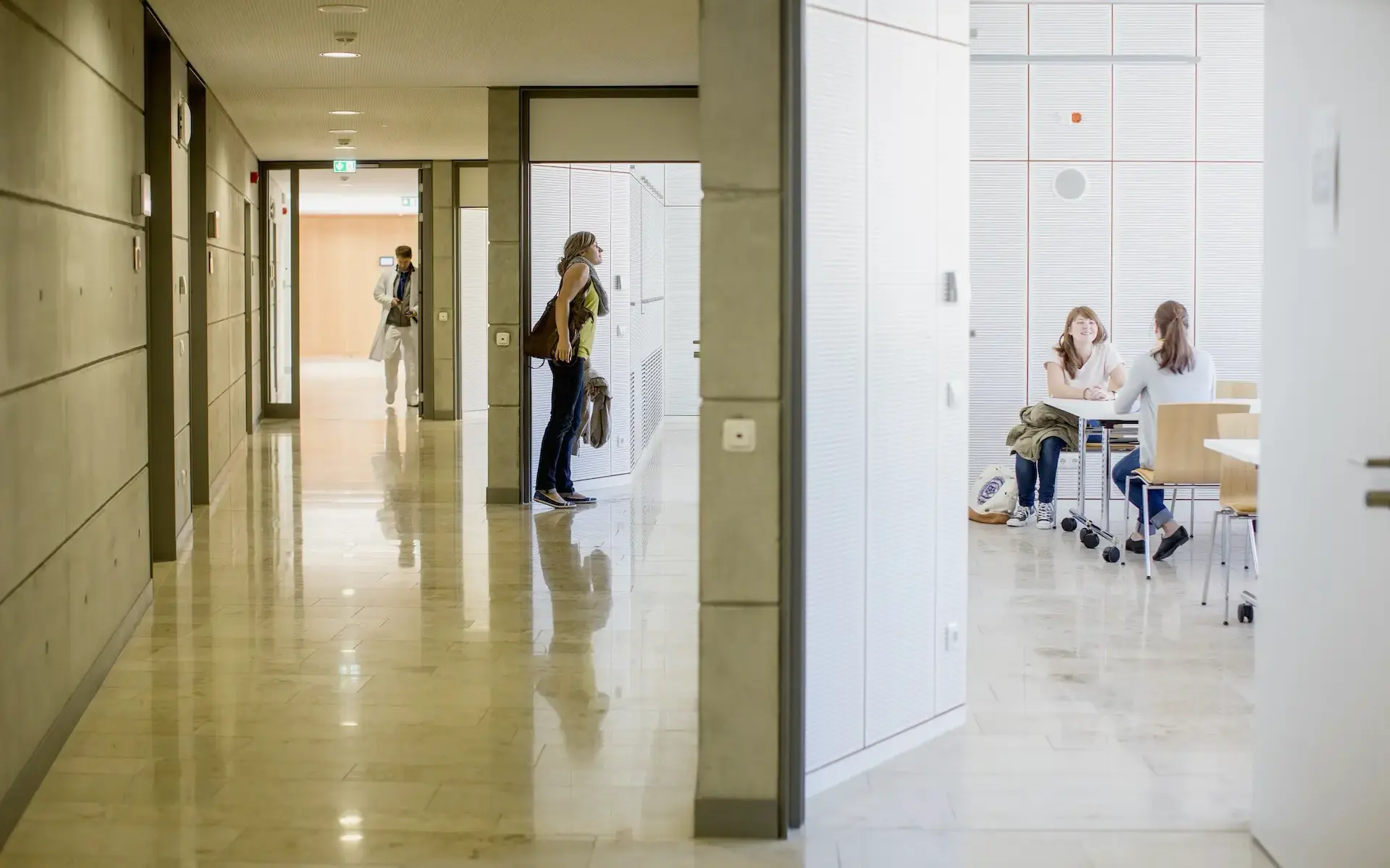Das Bild zeigt den Neubau des Lehr- und Lernzentrum in Essen. Abgebildet ist der zentrale Mittelflur, welcher das Gebäude längsseitig durchzieht, er ist gekennzeichnet durch Wände aus Sichtbeton, Muschelkalk auf dem Boden und einer Gipskarton-Abhangdecke.