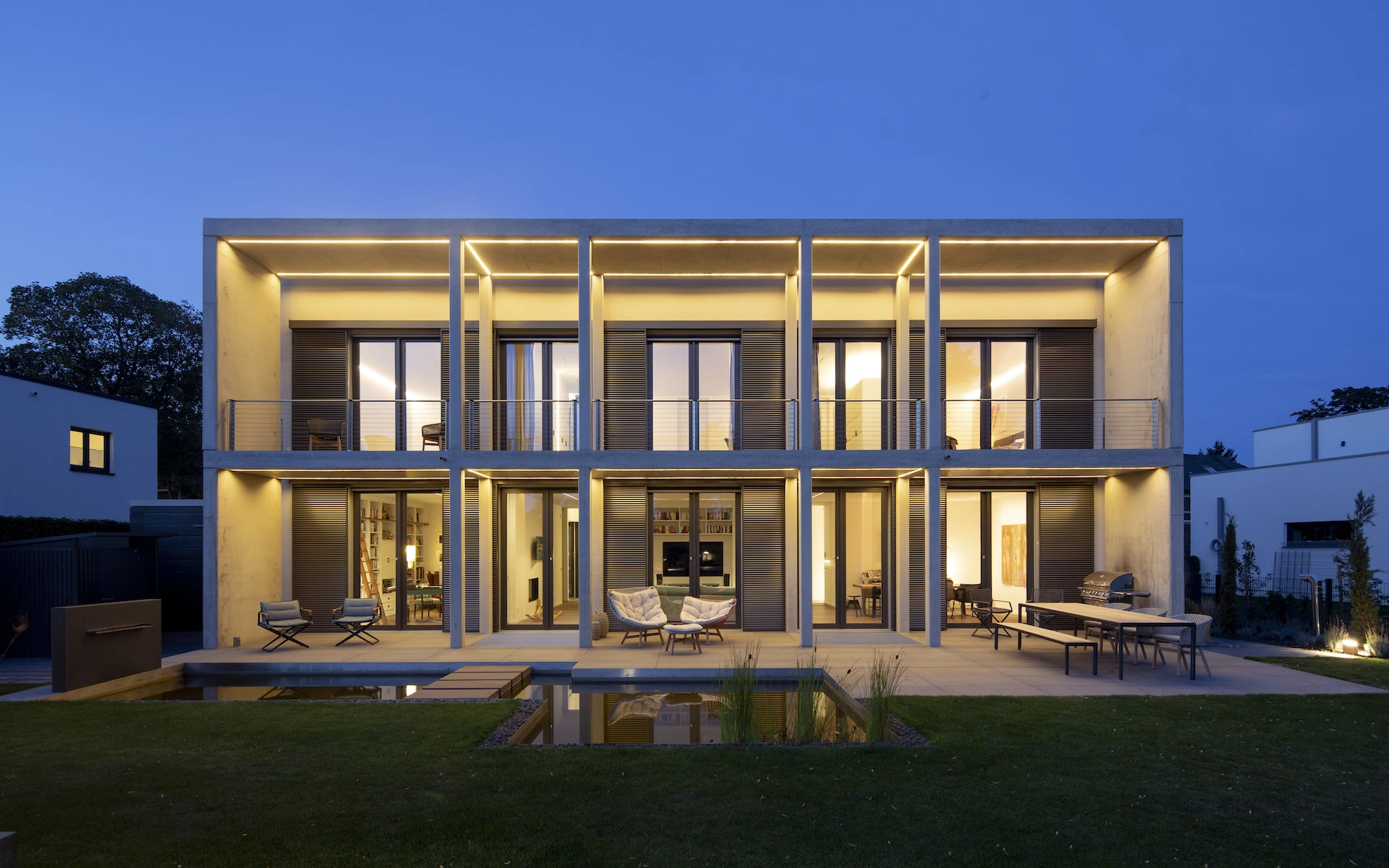 Neubau Haus K in Kamp-Lintfort Ansicht vom Garten in der Dämmerung mit raumhohen französischen Fenstern, elektrischen Schiebeläden, Sichtbetonveranda, linearen LED-Leuchten und reflecting pool.